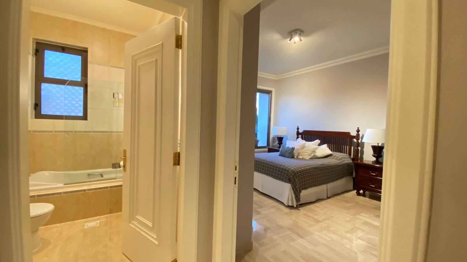 2346eb14-9bd2-4300-ad32-0fbd52172ddc View down a hallway with an open bathroom door to the left (bathtub visible) and a bedroom door to the right showing a wooden bed and two nightstands with lamps.