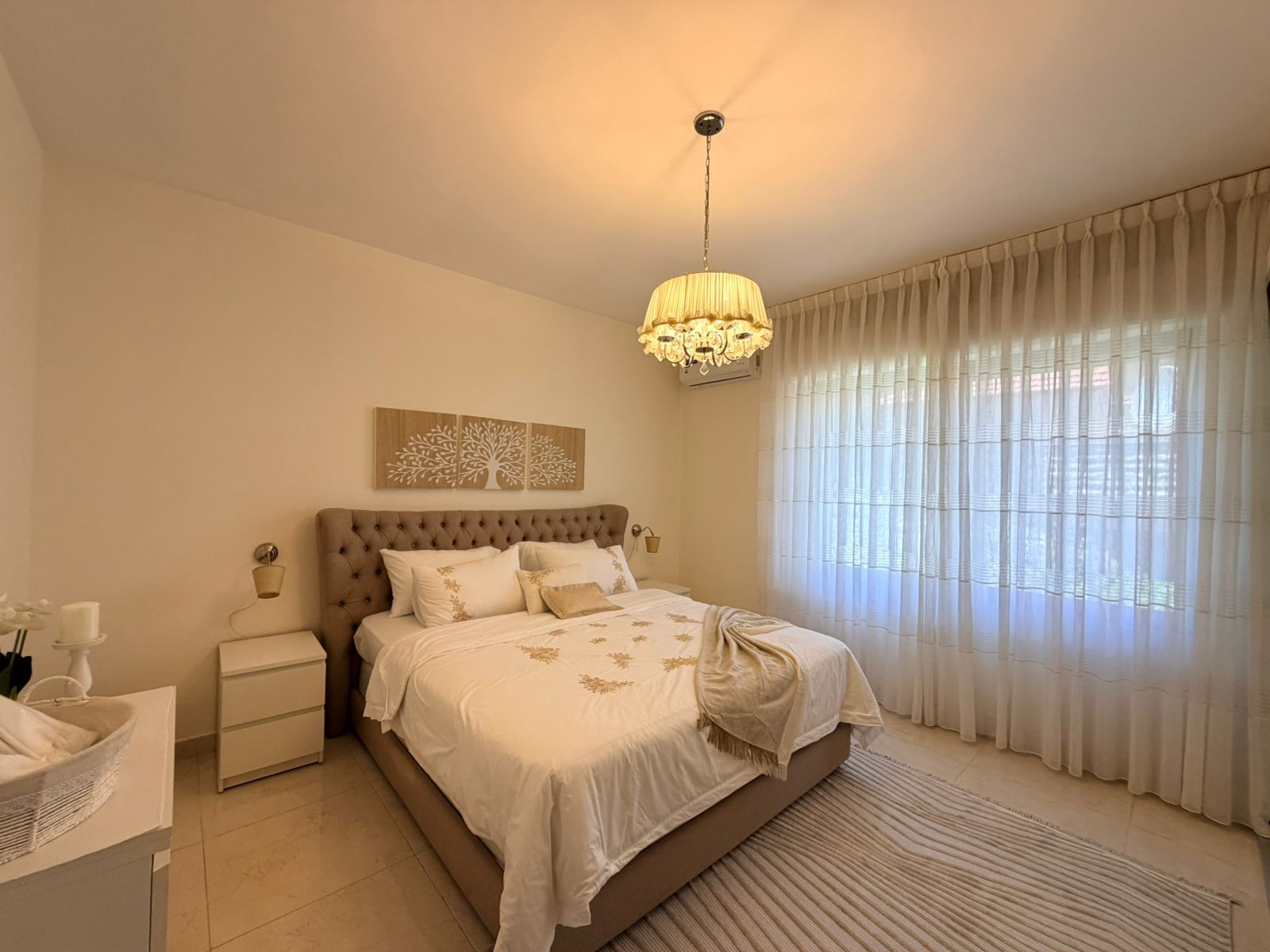 23483cfc-9f54-4155-afaa-4a49a69c6f88 Bedroom with a tufted beige headboard, white bedding with gold embroidery, and a chandelier overhead.