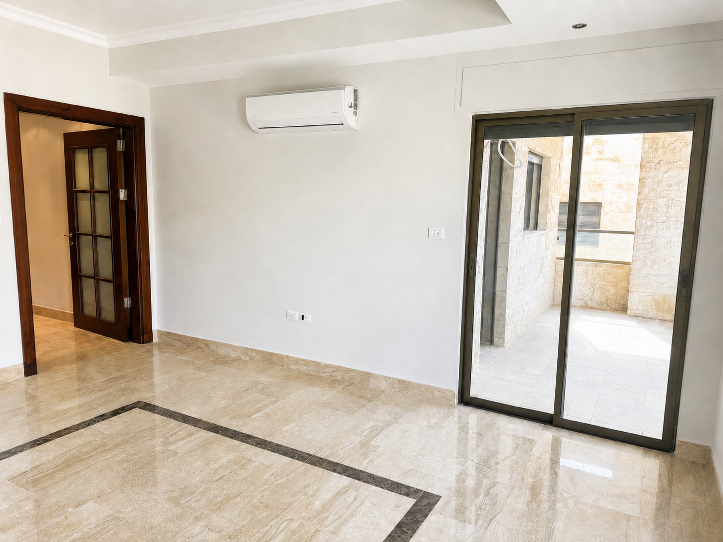 24343f5d-af2e-437c-85c7-01f61e260f82 1 Empty living room with beige marble tile, a wall-mounted AC, and a sliding glass door to an outdoor balcony.