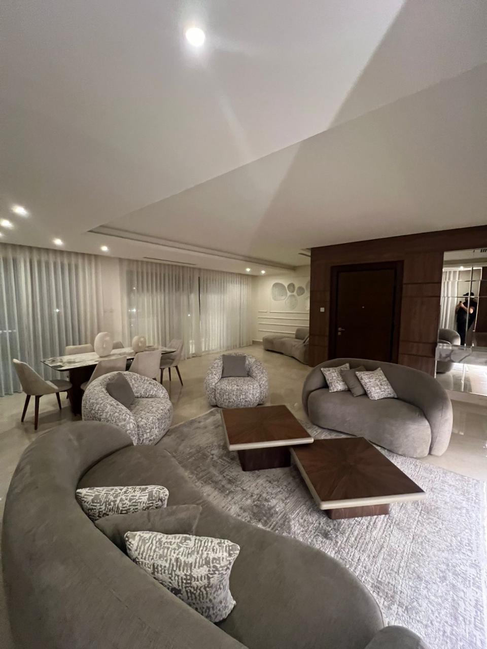 243d6b5a-0943-49c8-80bc-a5f91f1c2a9f Modern open-concept living room with curved gray sofas, patterned pillows, and two coffee tables on a light rug.