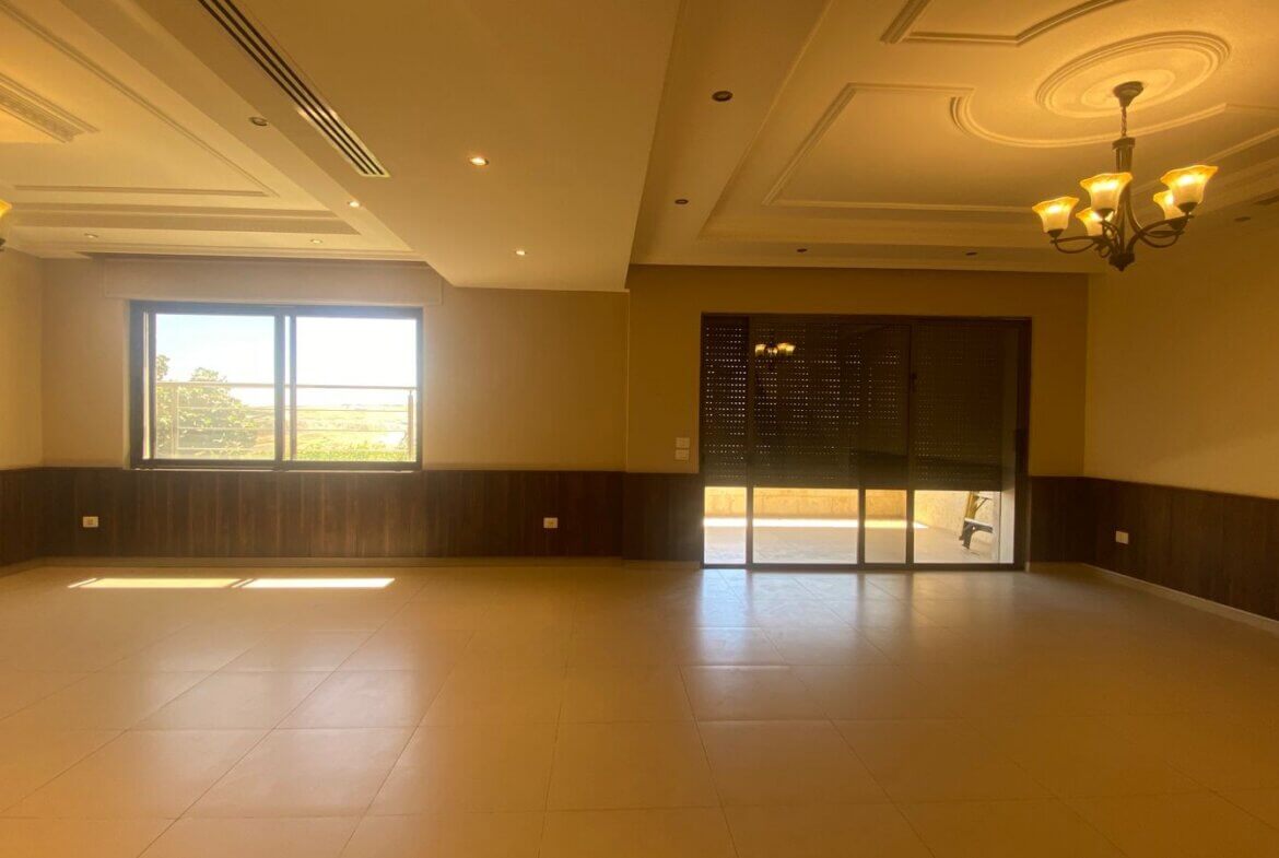 2456c201-bdb0-463f-a98d-e547f8a11707 Empty, spacious room with tiled floor, wood wainscoting, and two chandeliers; large window and glass door to the outside, letting in sunlight.