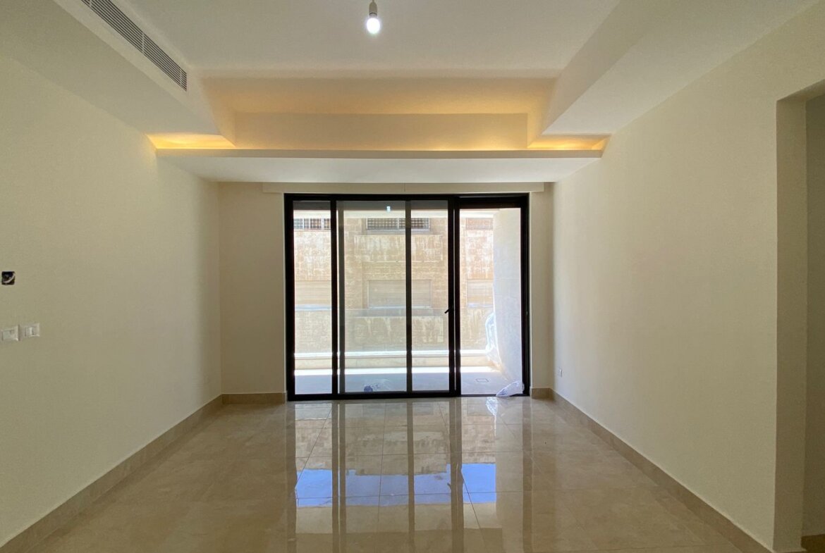 Bright empty living room with glossy beige tile floor and a large sliding glass door opening to a balcony; recessed ceiling lighting adds warm accents.