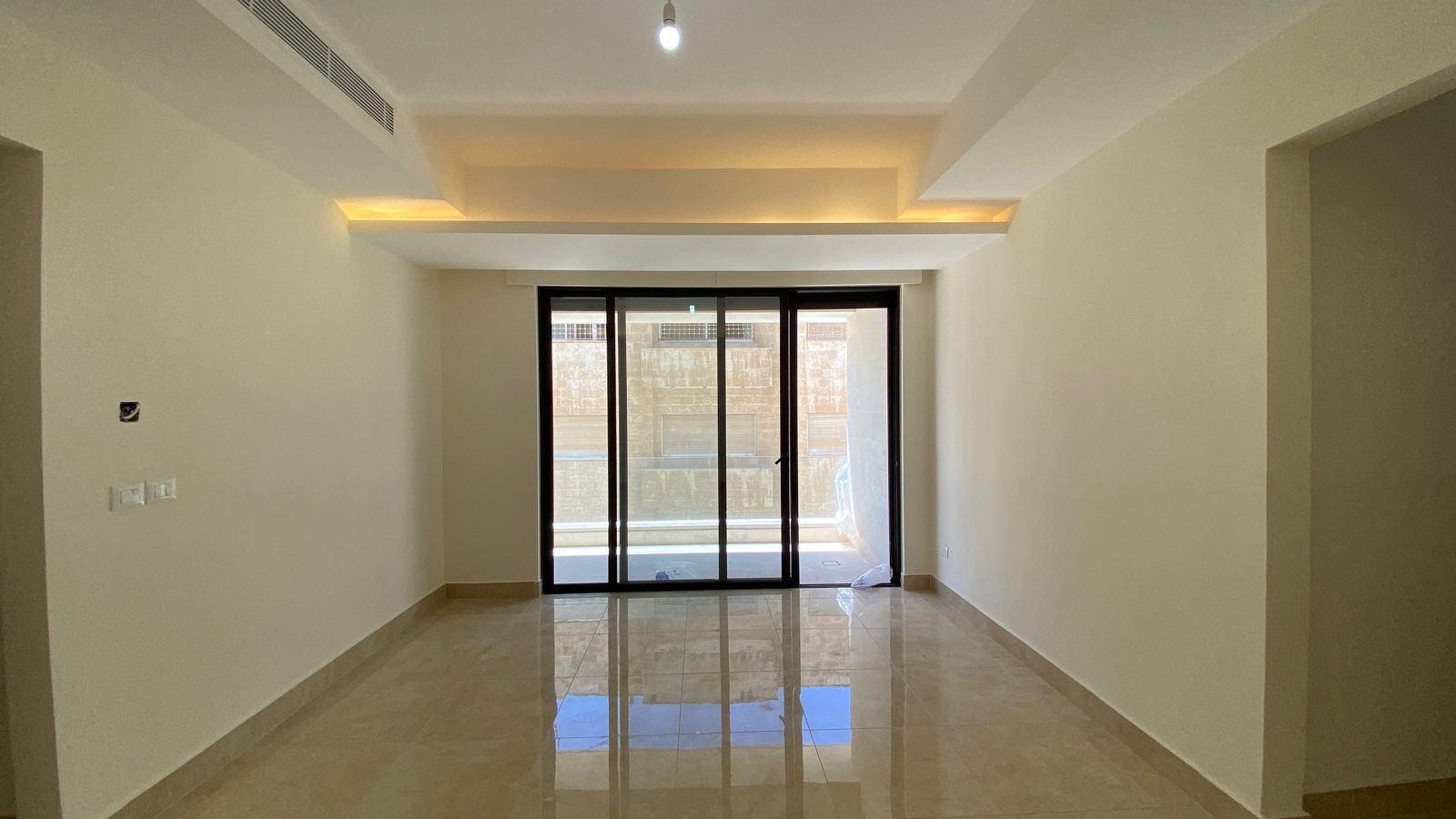 25173335-ddd2-44a7-8755-3fbae314e704 Bright empty living room with glossy beige tile floor and a large sliding glass door opening to a balcony; recessed ceiling lighting adds warm accents.