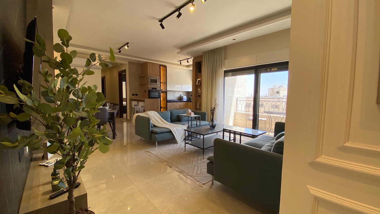 25aa1e2f-bd85-41d5-b411-575f2f69cb3d Open-concept living room with teal sofas, glass coffee table, and a balcony view through large sliding doors.