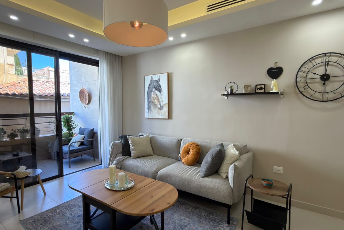 Modern living room with a gray sofa and colorful cushions near a sliding glass door to a balcony. A large horse portrait hangs on the wall, with a shelf and oversized wall clock nearby.