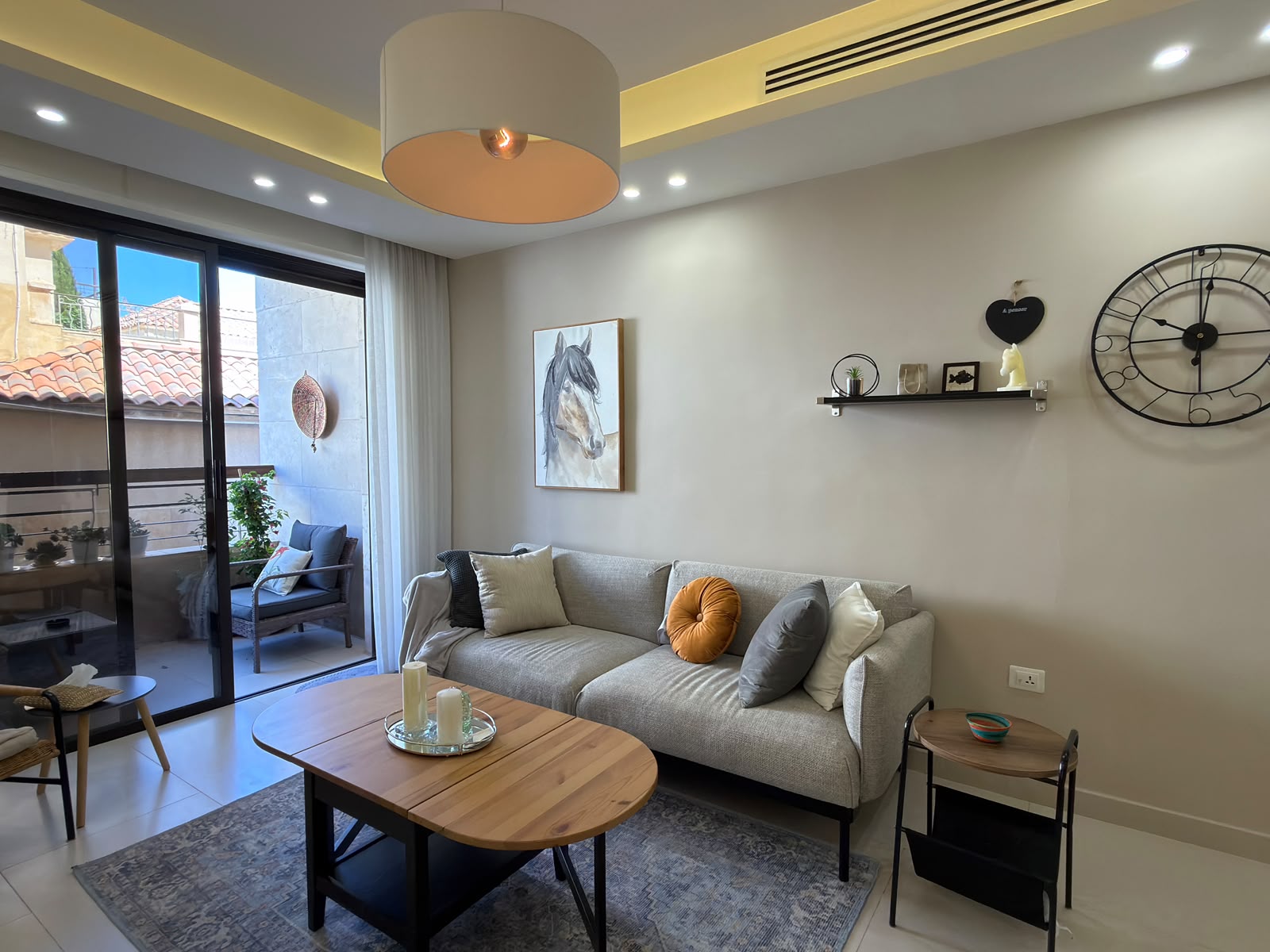 25cd2315-54df-4731-93b8-3399514de773 Modern living room with a gray sofa and colorful cushions near a sliding glass door to a balcony. A large horse portrait hangs on the wall, with a shelf and oversized wall clock nearby.