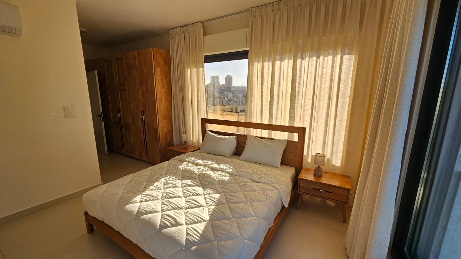 25eb9e1d-5137-4d46-aa65-81132dcc8109 Bright bedroom with a wooden bed, white quilt and two pillows, matching nightstands, and a large window with beige curtains showing a cityscape.