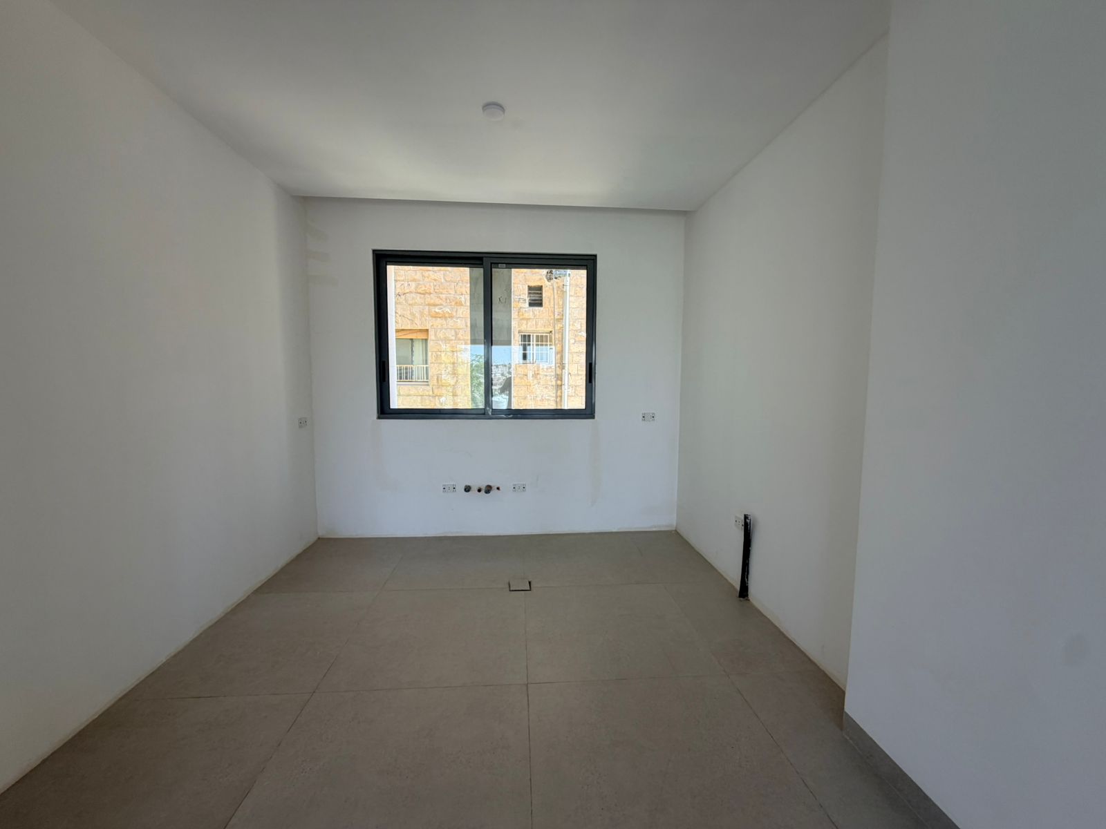 26a697b9-5415-4a24-bd69-4246e9dbf2b9 A small unfurnished white room with a single window centered on the far wall, beige floor tiles, and exposed outlets along the base of the wall beneath the window; appears to be under construction or renovation.