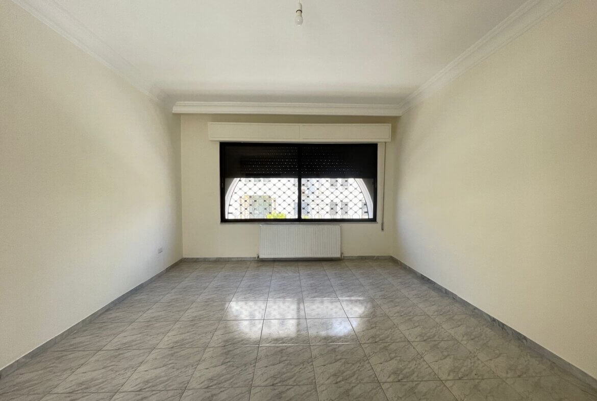26e1b466-b546-4070-b68c-3a5840ebcc0e Empty living room with a large barred window, radiator beneath, and pale walls with tiled floor.