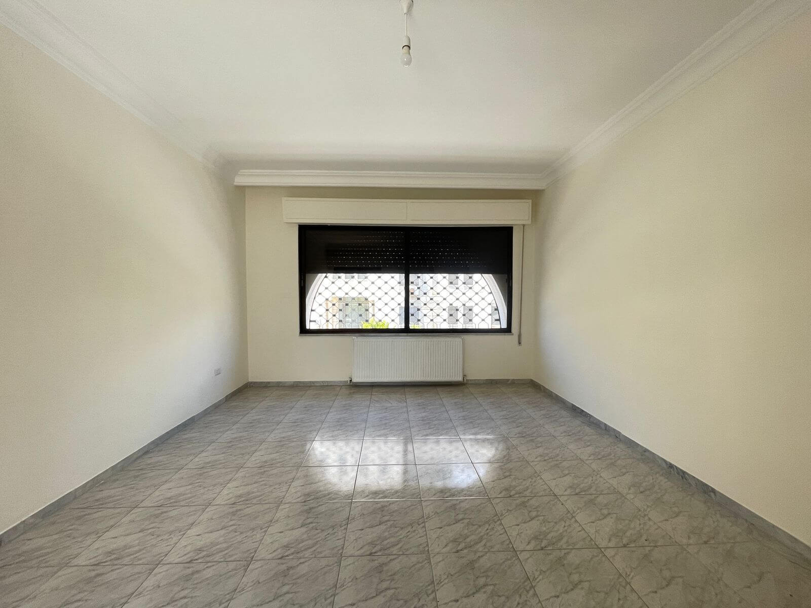26e1b466-b546-4070-b68c-3a5840ebcc0e Empty living room with a large barred window, radiator beneath, and pale walls with tiled floor.