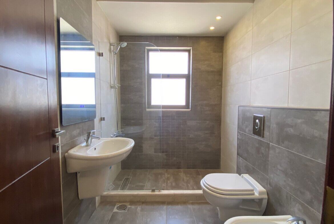 27972e38-f192-4029-a4e7-f985f861769b Modern bathroom with a walk-in shower, white pedestal sink, and wall-hung toilet in gray tiled room; small window above the shower and recessed ceiling lights illuminate the space.