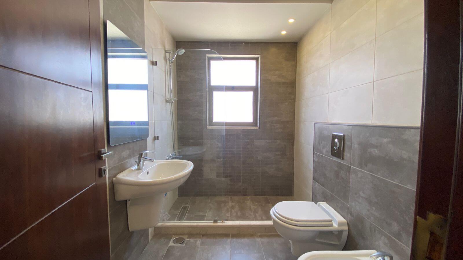 27972e38-f192-4029-a4e7-f985f861769b Modern bathroom with a walk-in shower, white pedestal sink, and wall-hung toilet in gray tiled room; small window above the shower and recessed ceiling lights illuminate the space.