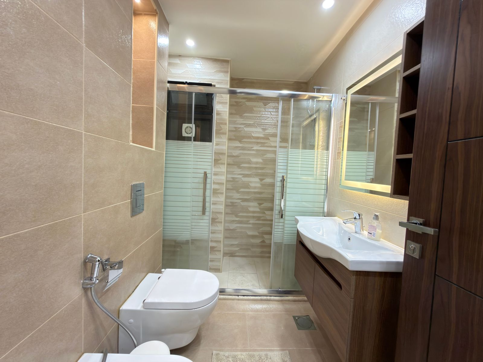 27e5895e-17e9-430d-83fa-66f9bd20ee6b Modern bathroom with glass shower enclosure, white sink, and wooden vanity along a beige-tiled wall.