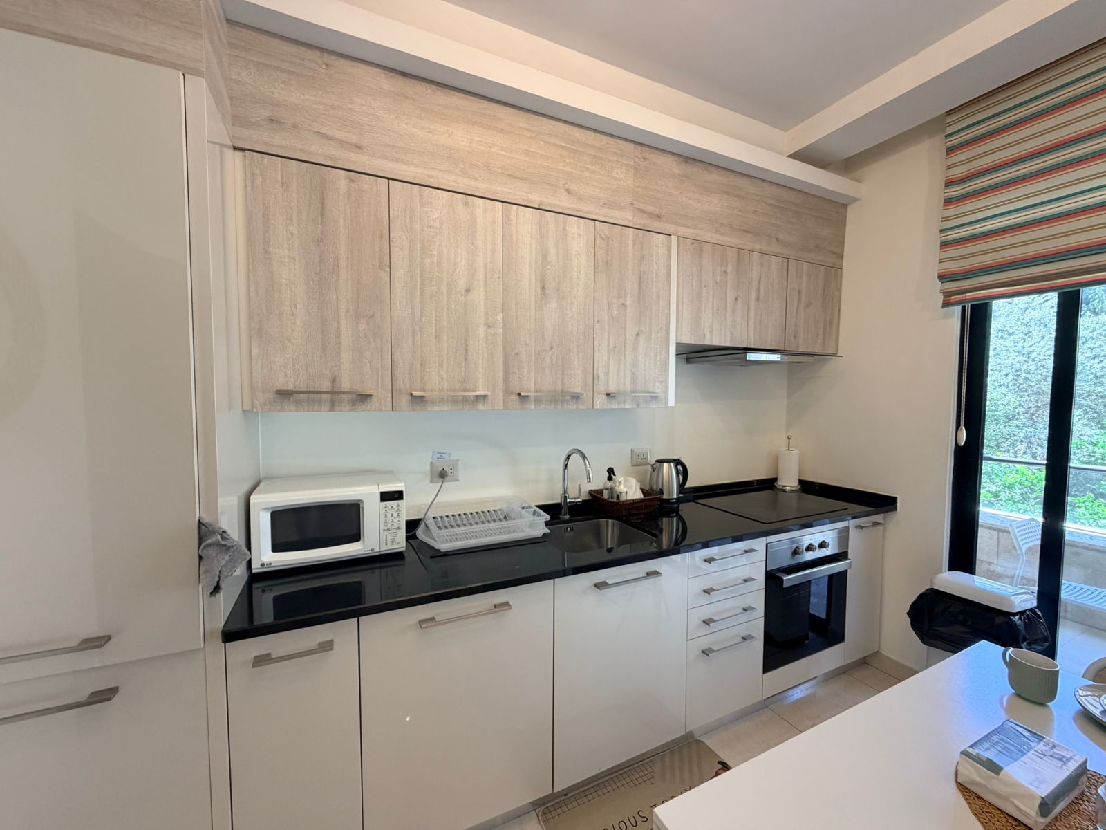27f1e82a-c9fb-4c76-867e-74548fea04a5 Modern kitchen with white base cabinets, light-wood upper cabinets, and a black countertop, featuring a sink, dish rack, microwave, and kettle.