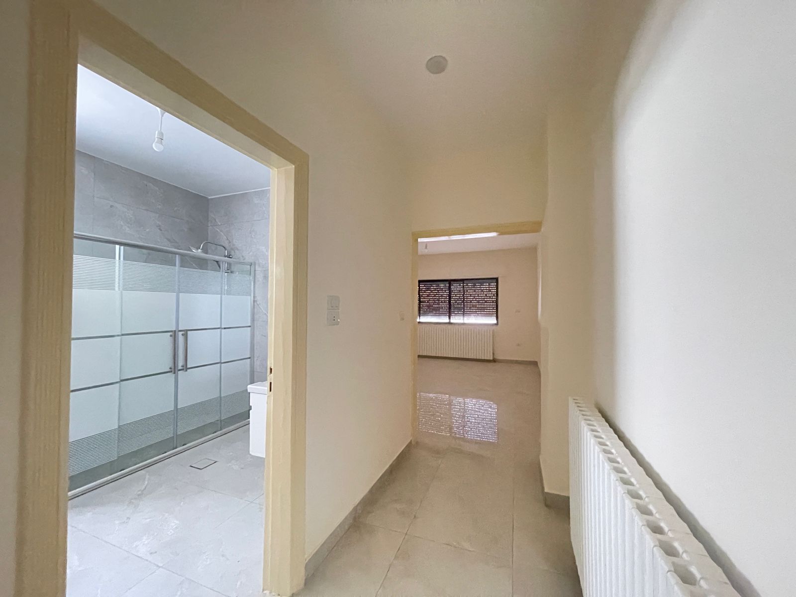 29d9c510-905a-47f7-a052-7da1301d6466 Hallway view with a bathroom on the left featuring a frosted glass shower enclosure, beige walls, and a radiator along the right wall. A window with blinds is visible at the far end.
