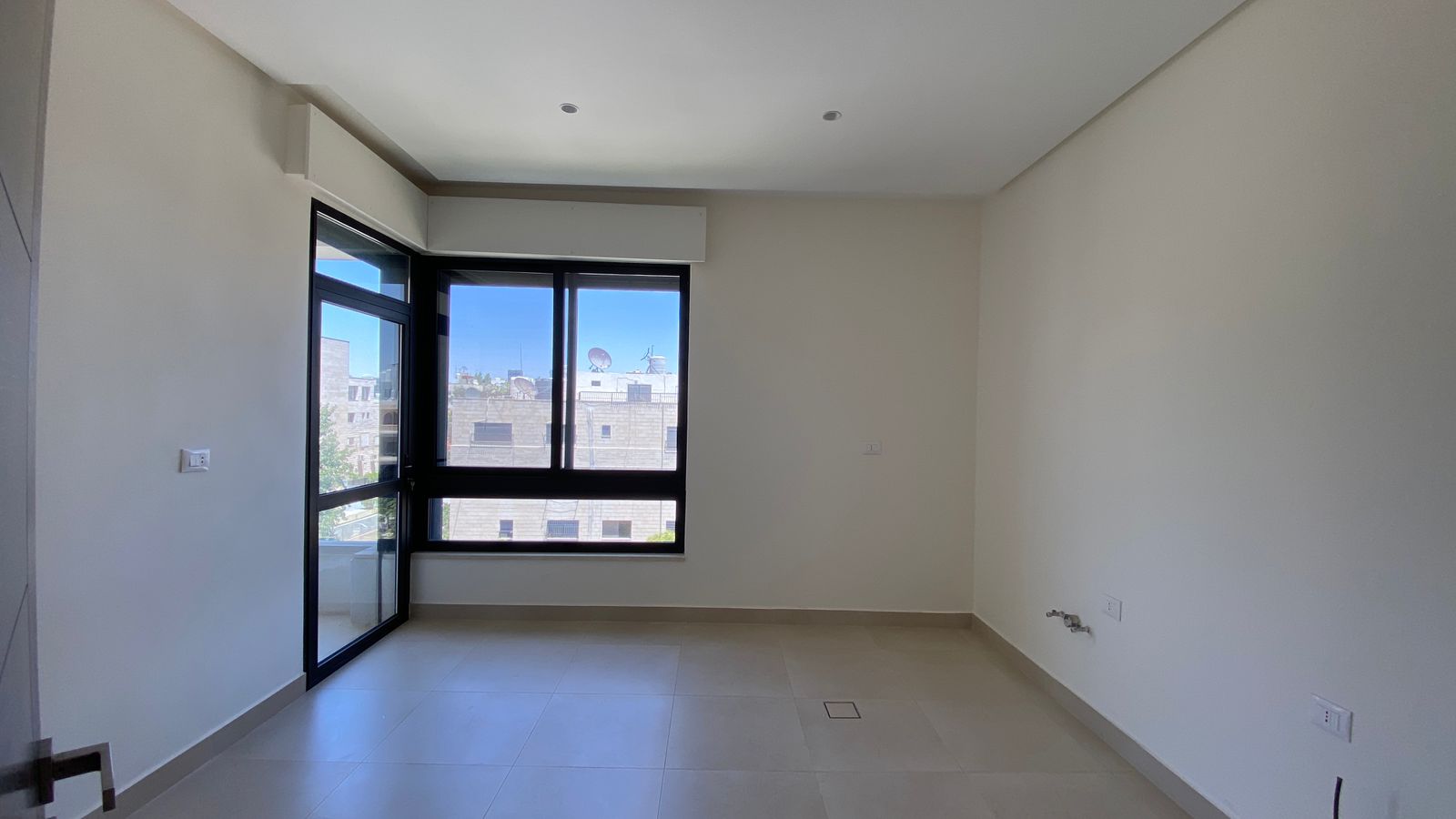 2a28b2d8-f60e-4063-a595-2df510ed51ee Unfurnished room with light beige tile floor, white walls, and a large black-framed window and balcony door; city buildings visible outside.
