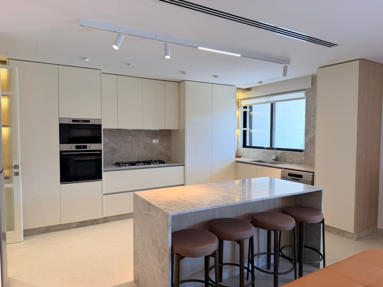 2a535d25-c68b-4e18-be1e-13dae4212f85 Modern white kitchen with built-in double ovens, gas cooktop, marble island, and row of brown bar stools.