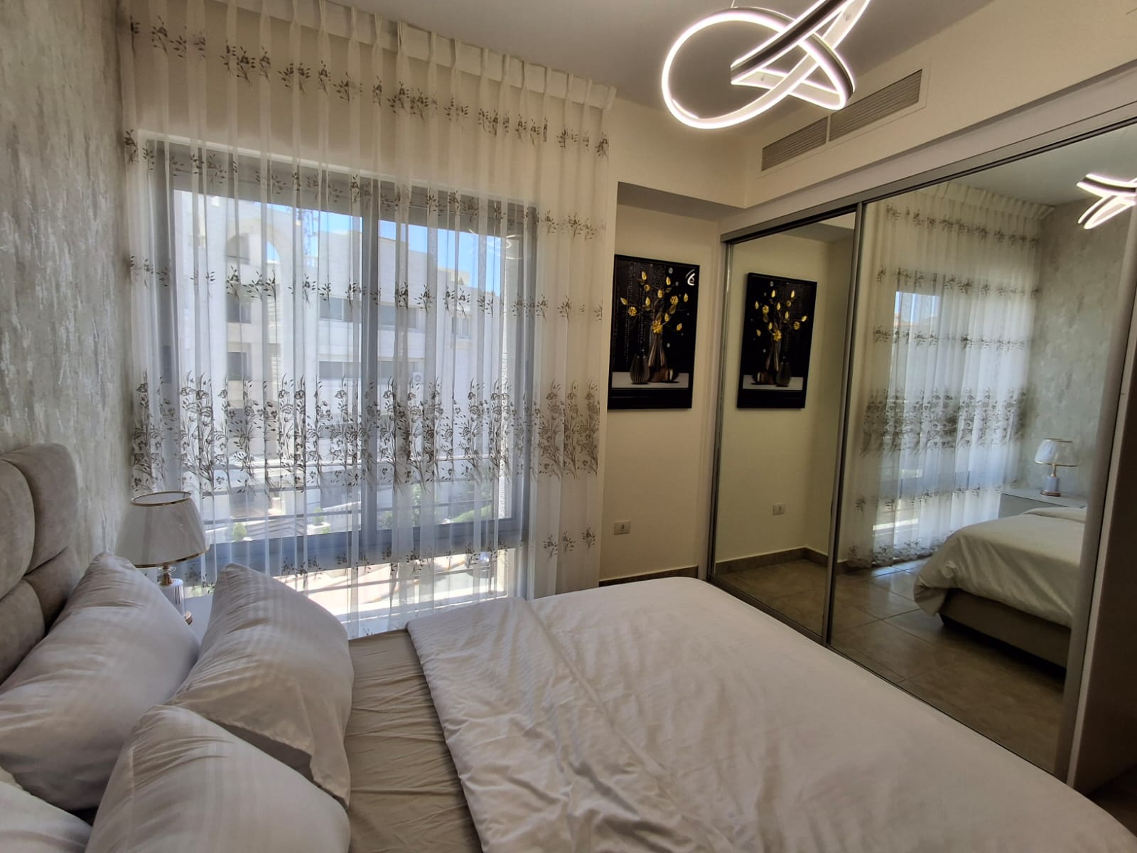 2ac33854-5ca4-41fb-a030-fb394d7e8dc7 Bright modern bedroom with a white bed, sheer floral curtains, and a large window letting in daylight. A mirrored sliding wardrobe reflects the room, and a contemporary circular ceiling light hangs overhead.