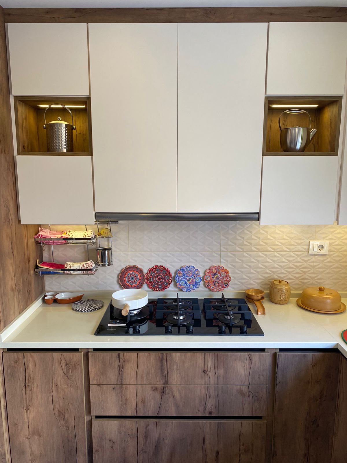 2b090f97-f4b2-4b39-8112-0334dd00a3a6 Modern kitchen with white upper cabinets, wooden lower cabinets, and a gas stove; decorative plates on the backsplash and kettles in shelves.