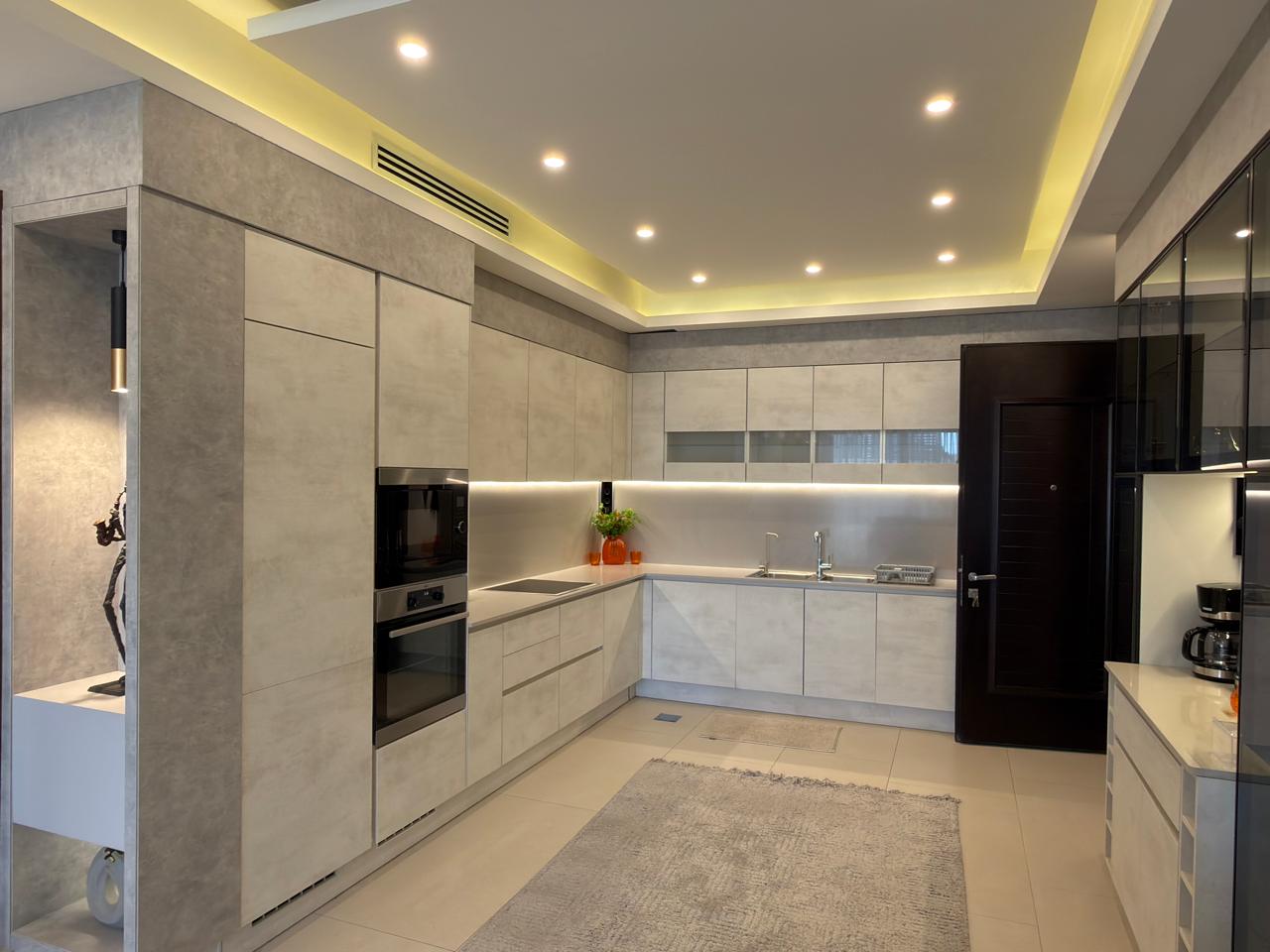 2b5b5b66-1145-4bdb-adc4-b65e51ab970b Modern L-shaped kitchen with light gray cabinets, built-in oven/microwave, and under-cabinet lighting along a glossy backsplash.