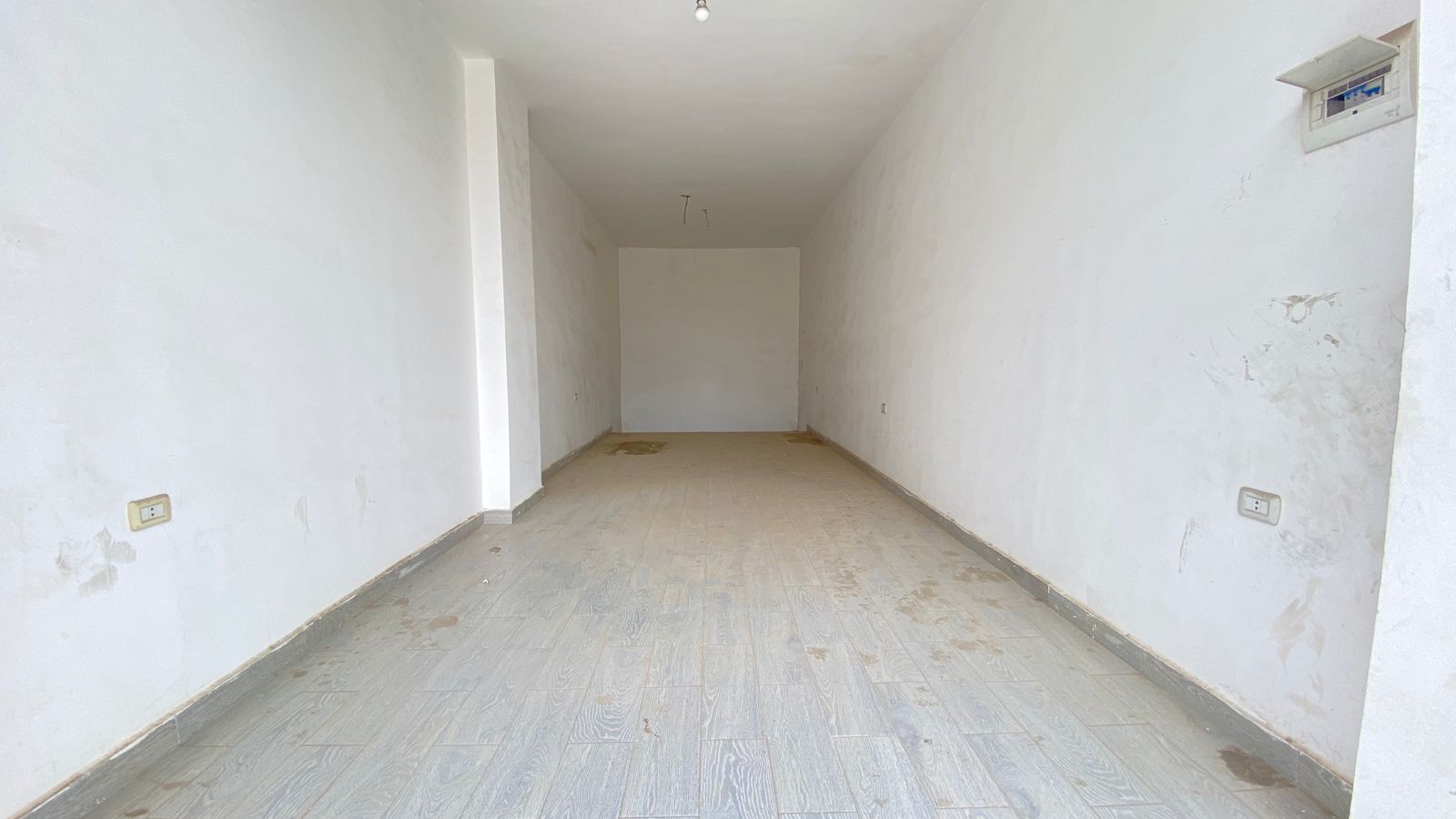 2ba72227-645a-470f-b4d2-46ef3999bd55 Long, empty white-walled room with a tiled floor, seen from the entrance, and a bare light bulb hanging from the ceiling.