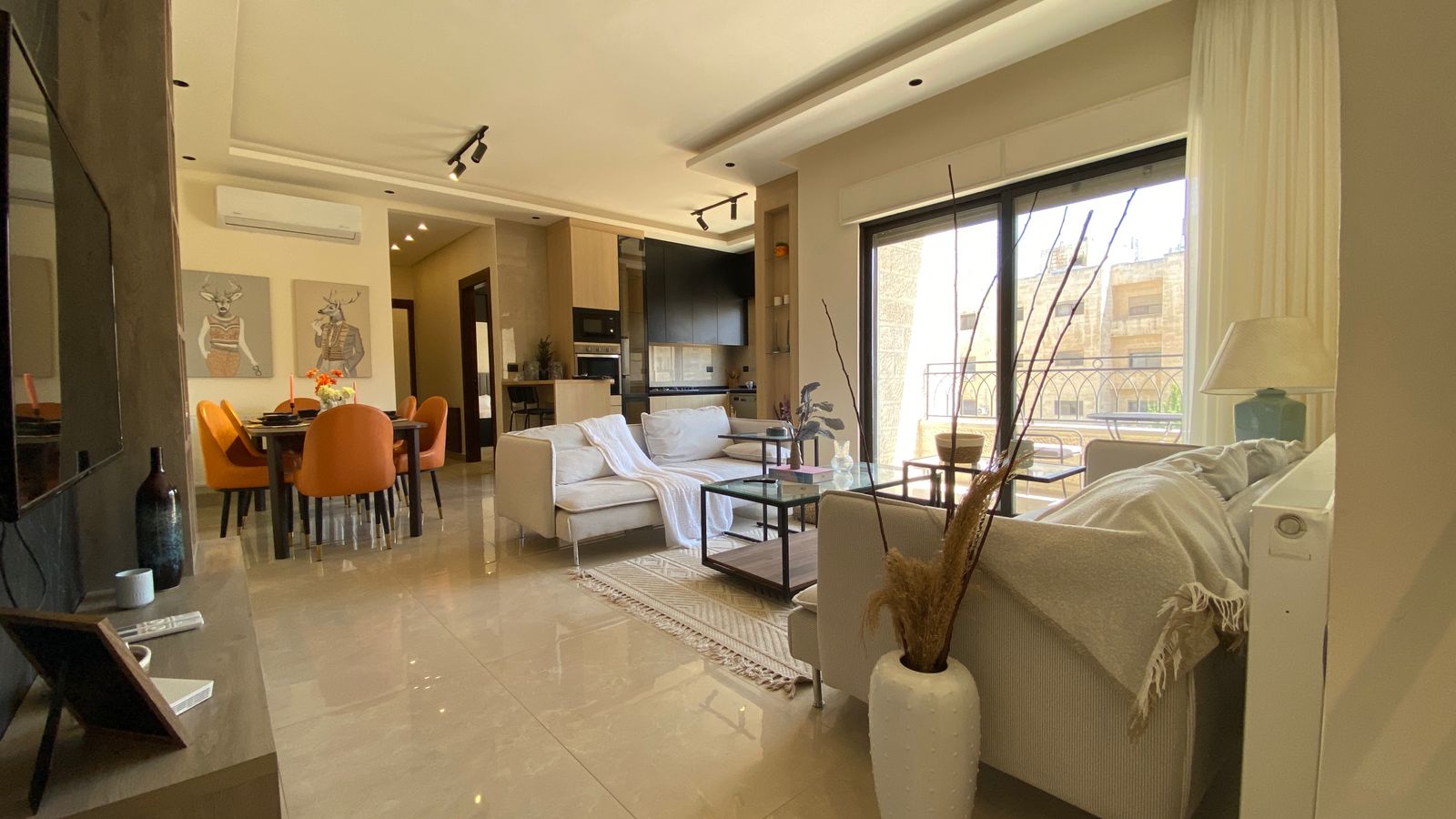 2be80566-7289-4084-ba7e-5b336ed4d580 Open-concept living and dining area with a cream sofa, glass coffee table, and bright orange dining chairs near a large window. In the background is a kitchen.