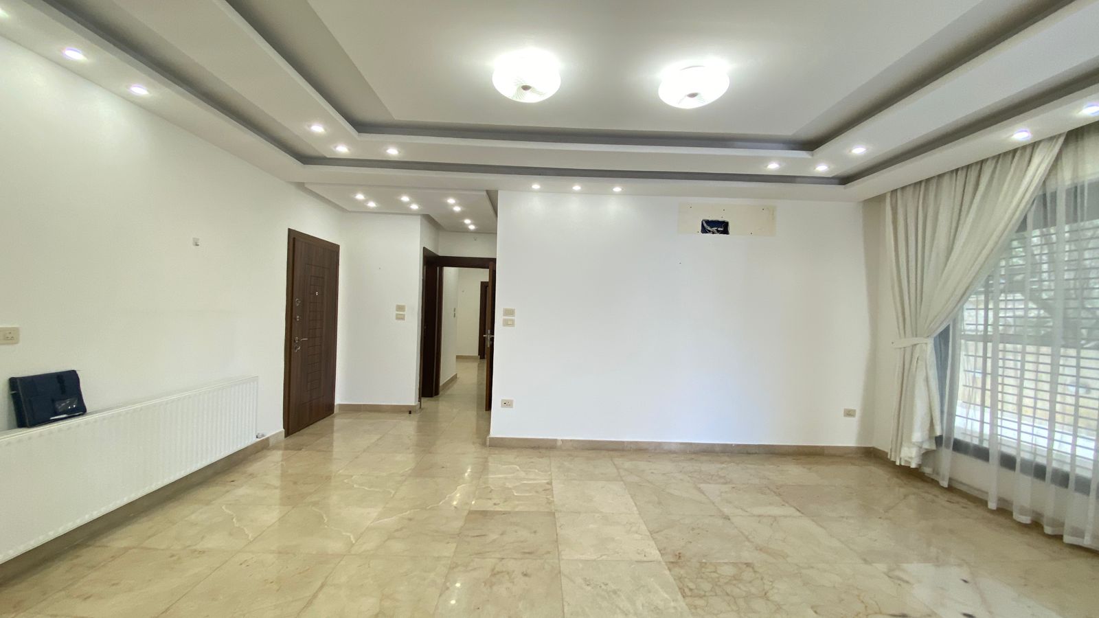 2c266314-37a0-47b0-bfc5-393f203d690d Bright empty living room with marble floor, white walls, recessed ceiling lights, a dark wooden door, and a large window with sheer curtains.