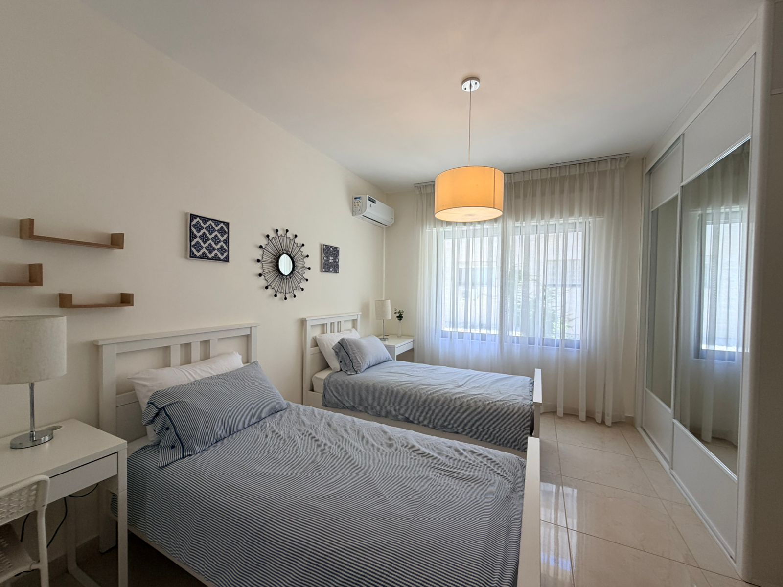 2c5d51db-ccec-414e-a53b-bfd1e62ff767 Twin bedroom with two white-frame beds and blue striped bedding, minimalist decor, and a sunburst mirror on the wall above.