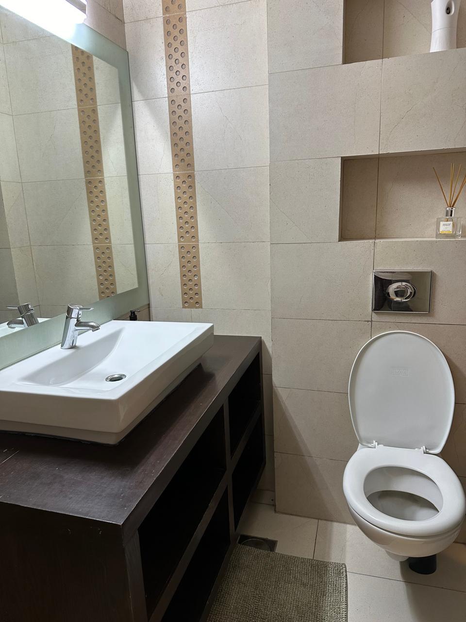 2d6e5725-f6b3-410b-847b-5eb6d75214f9 Bathroom sink with white rectangular basin and chrome faucet against a dark wood vanity with open shelves, mirror above, and beige tiled walls.