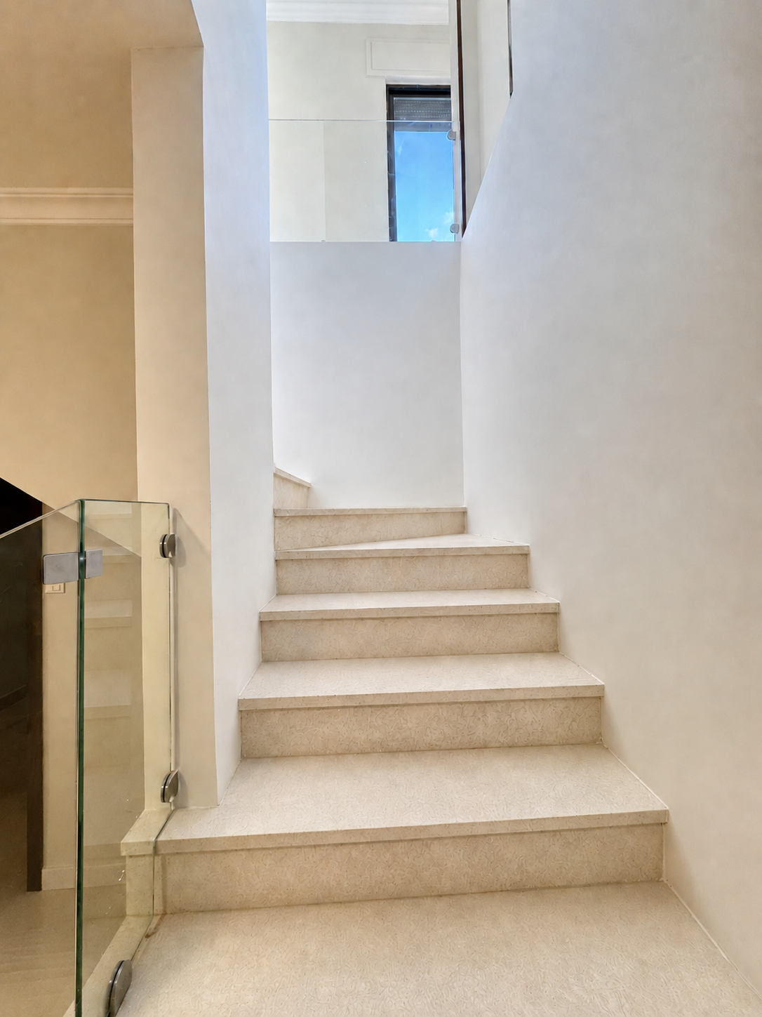 2ddd3b9d-785c-48fd-9bd9-8e198679919c Interior staircase with beige stone steps and glass railing on the left, leading up to a bright landing with a window.