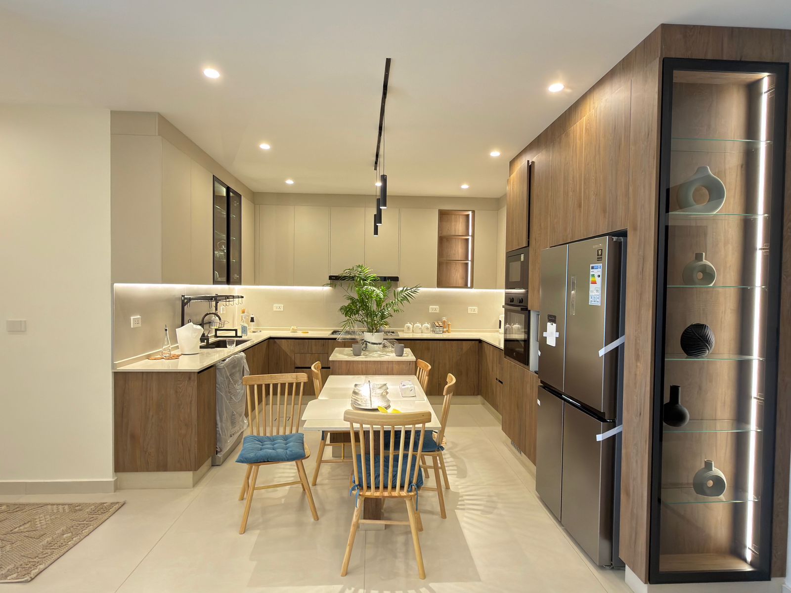 2e0efc1c-0fd0-4e9b-897c-f3095f0d6aca Open-plan kitchen with wood-tone cabinets, stainless steel fridge, glass display cabinet, and a four-seat wooden dining table with blue cushions in the foreground.