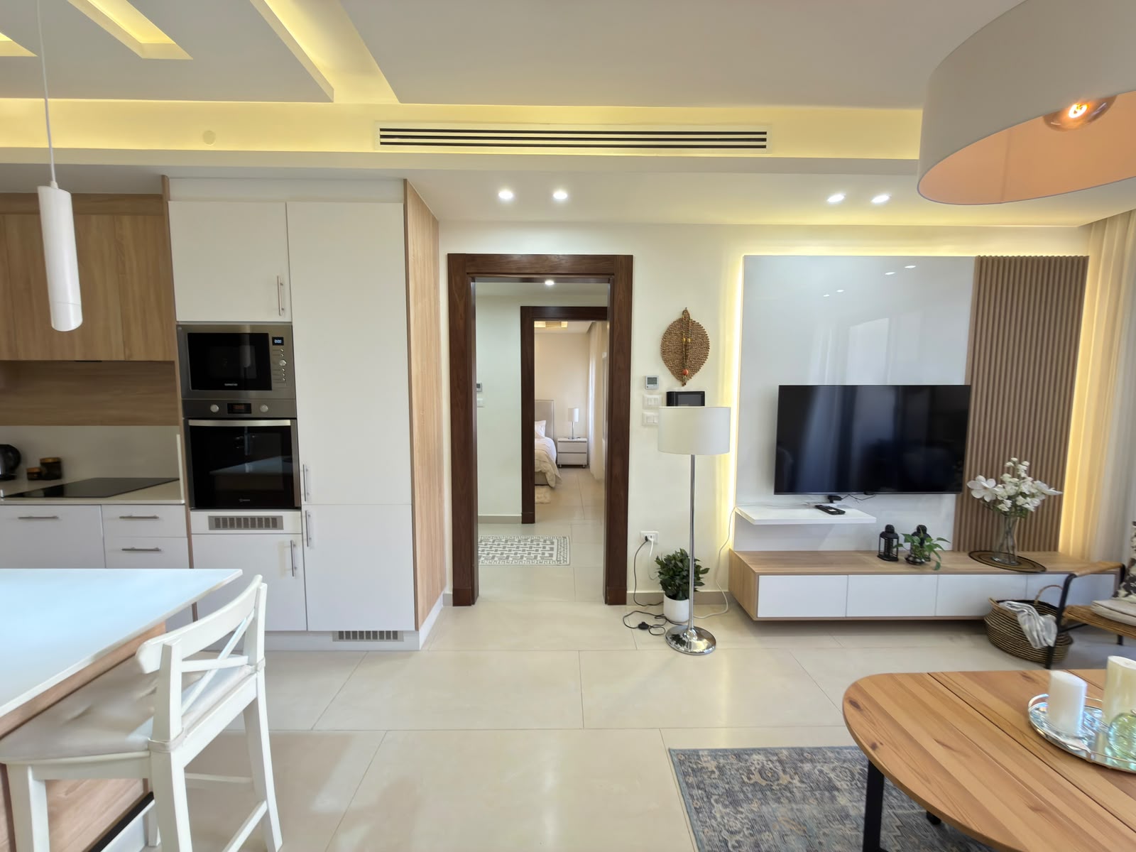 30c9a9f0-9b23-4a96-b960-12574b605a92 Bright open-plan living space with a white-and-wood kitchen on the left, a doorway to a bedroom in the center, and a TV wall on the right.