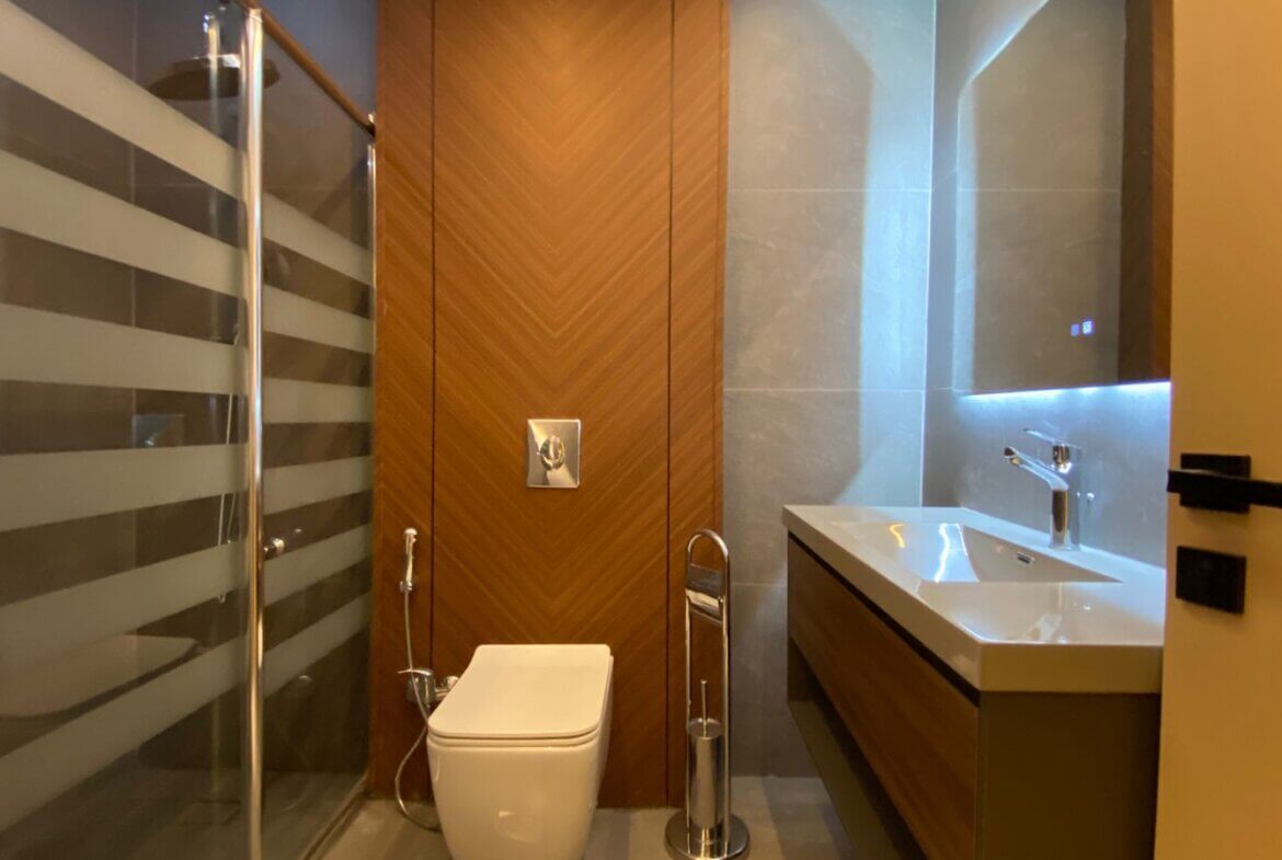 Modern bathroom with a wall-mounted toilet, wooden chevron accent wall, glass shower door, and illuminated vanity mirror.