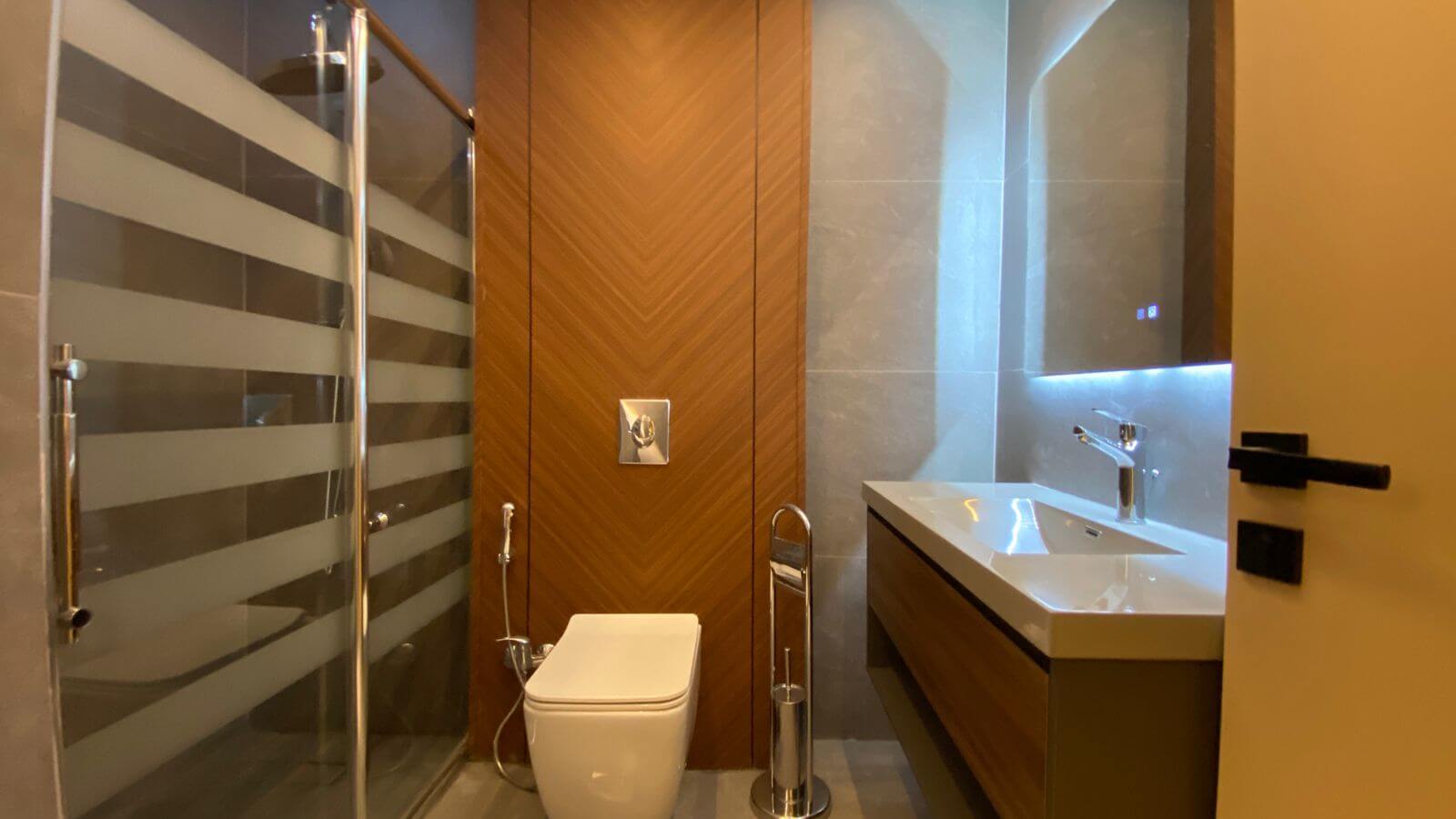 Modern bathroom with a wall-mounted toilet, wooden chevron accent wall, glass shower door, and illuminated vanity mirror.