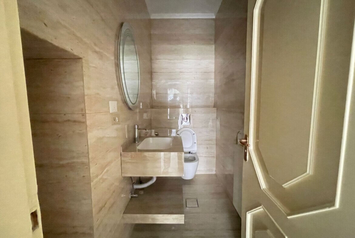 3311c8fe-7a4e-4b82-b11c-685833f4ba95 Modern beige-tiled bathroom with a round wall mirror, sink vanity, and toilet.