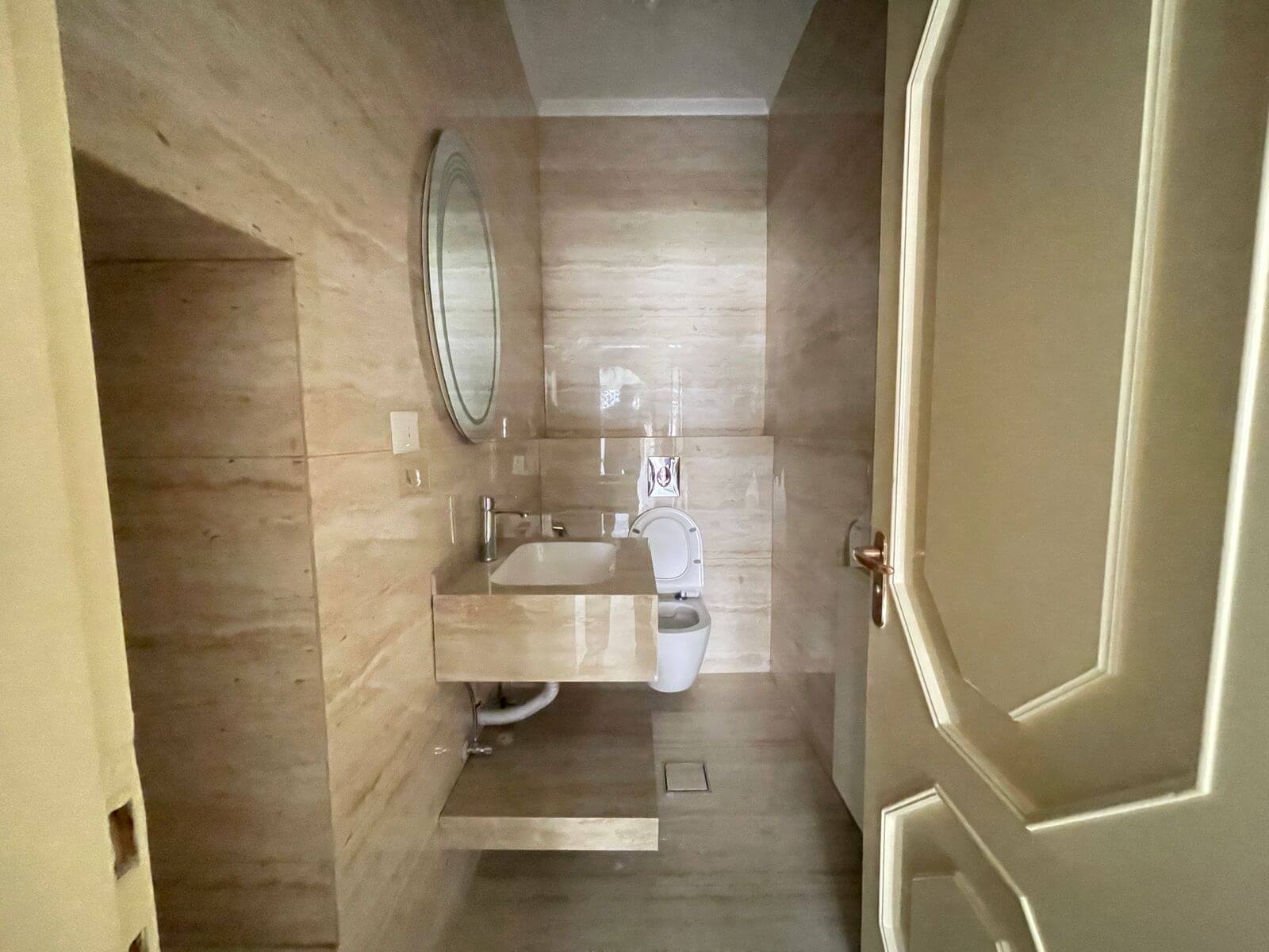 3311c8fe-7a4e-4b82-b11c-685833f4ba95 Modern beige-tiled bathroom with a round wall mirror, sink vanity, and toilet.