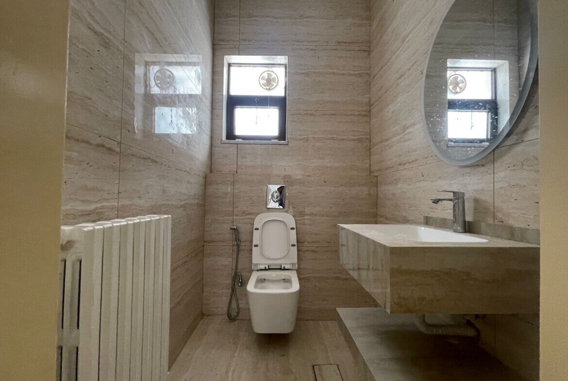 331f22b2-e6a6-4bea-b79b-7cfe2f6bbb11 Marble bathroom with wall-mounted sink, round mirror, and a toilet facing a small window; beige stone walls.