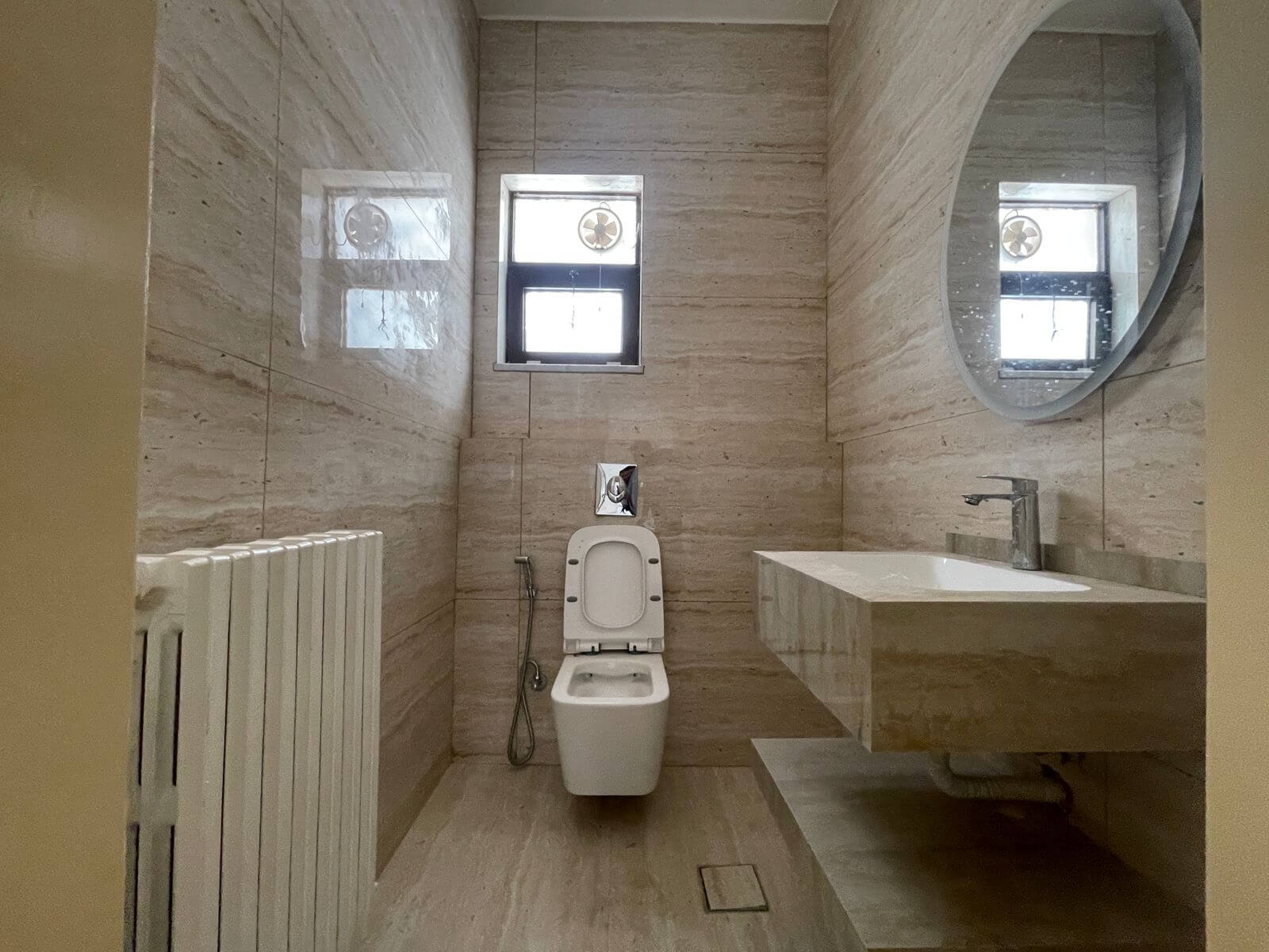 331f22b2-e6a6-4bea-b79b-7cfe2f6bbb11 Marble bathroom with wall-mounted sink, round mirror, and a toilet facing a small window; beige stone walls.