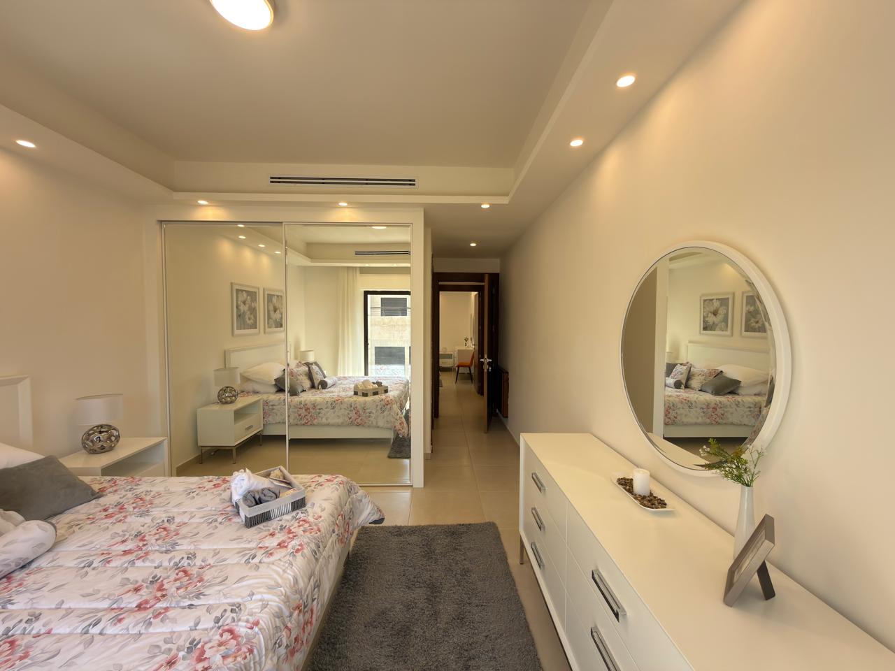 333ee2ee-724b-4bca-b6c6-c0426765e0c9 Bright bedroom with a floral quilt on the bed, white nightstands, and a large white dresser with a round mirror above it.