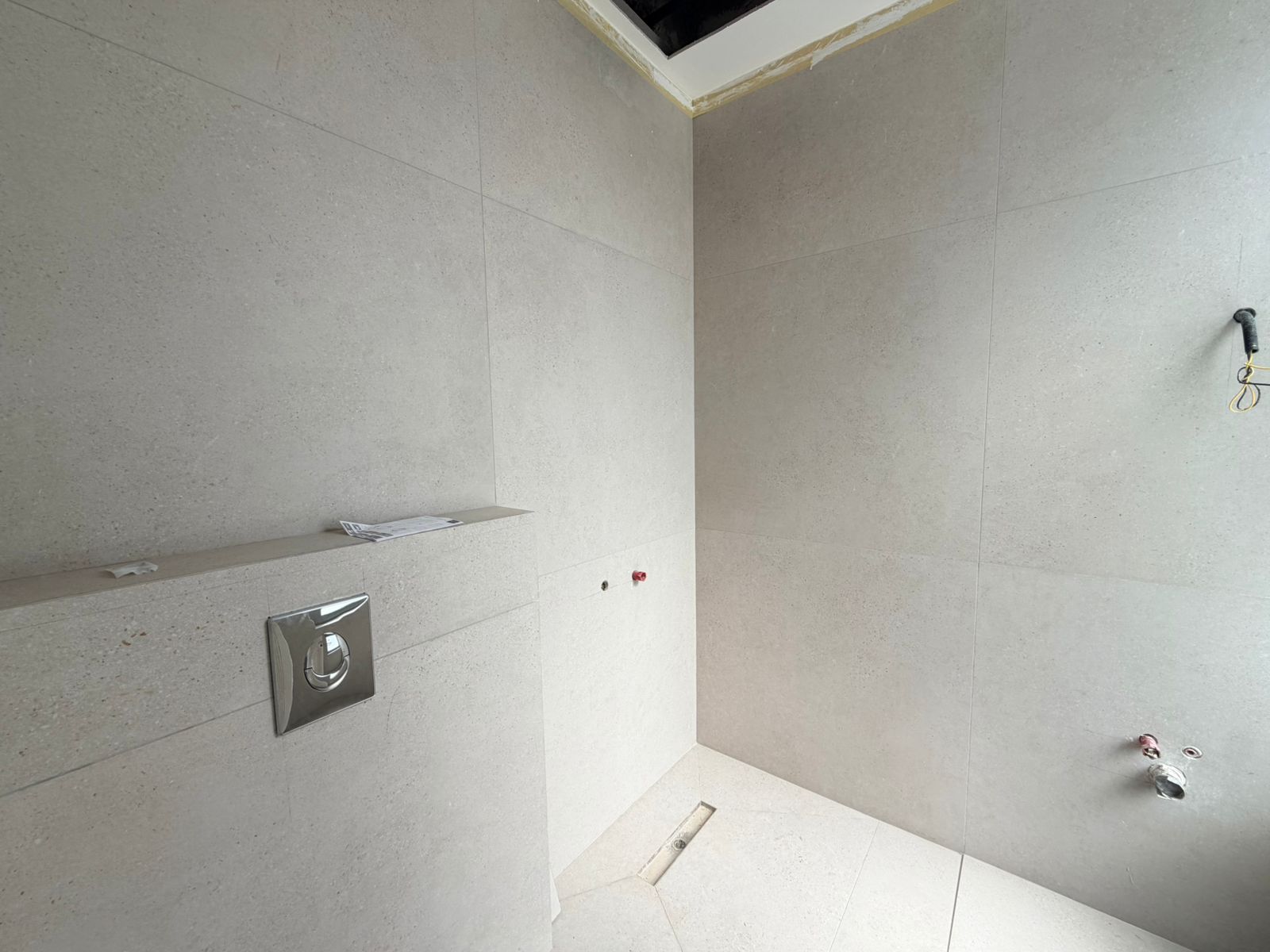 346251c1-b252-49bf-bb5b-12f7570f735d Under-construction bathroom with gray tiled walls, a wall-mounted flush plate, open ceiling hatch, and exposed wiring on the right side.