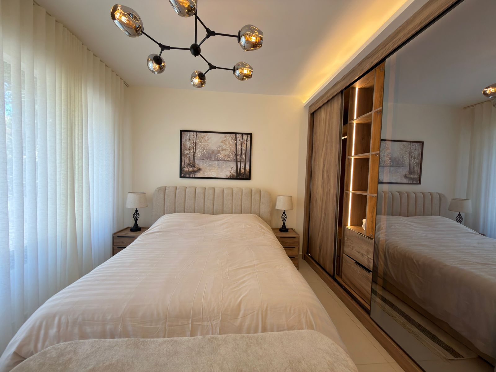 34714e06-3f08-4ce7-a290-925eebbceb57 Bright modern bedroom with a large bed and beige upholstered headboard, two nightstands with lamps, a framed landscape painting, and a mirrored wardrobe with warm lighting.