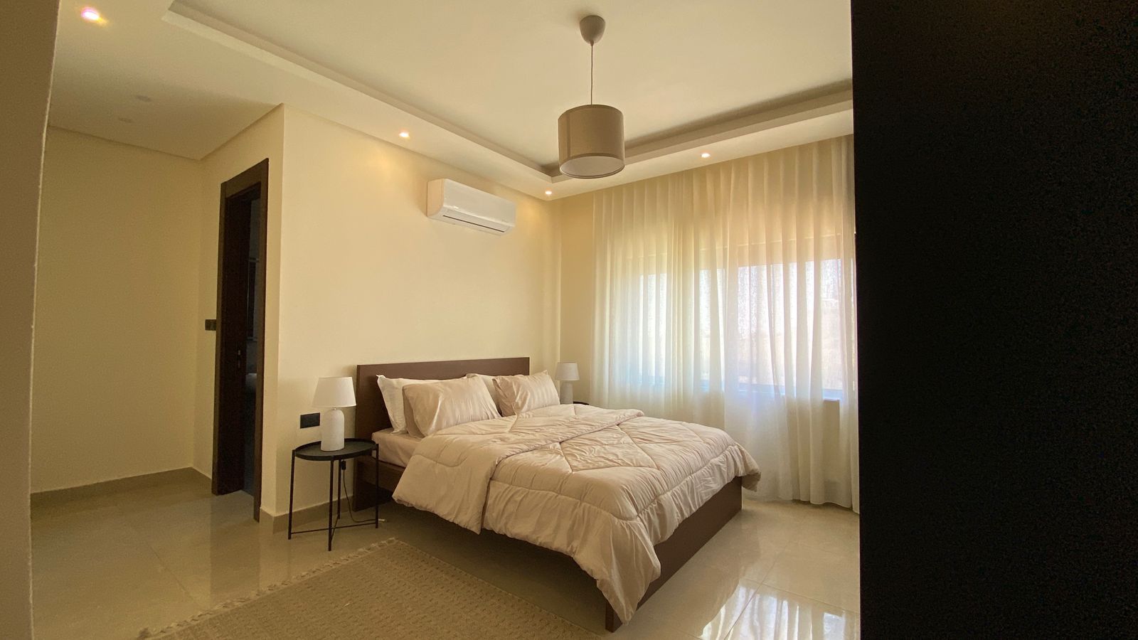 34dd9f8d-60f4-41f7-b659-3adeb2cf49e0 Modern hotel-style bedroom with a queen bed, white comforter, two nightstands with lamps, and sheer-curtained window in a bright room.