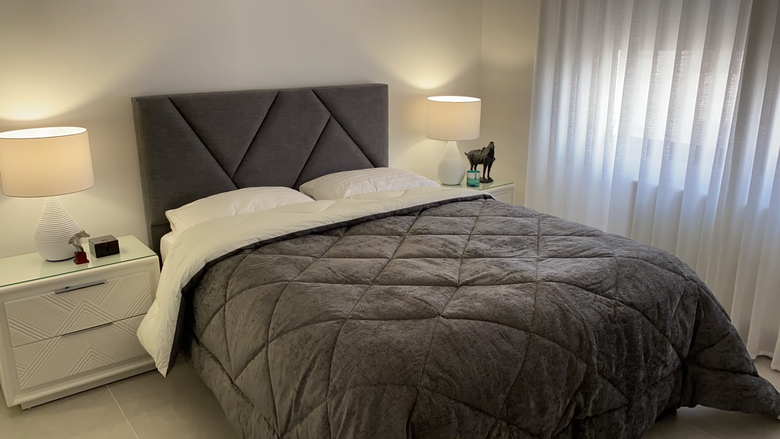 34e48fa3-46eb-4b65-bd74-ae25d4d78d39 Modern gray upholstered bed with a diamond-tufted headboard, white pillows, and a dark gray quilt in a bright, minimal bedroom setting.