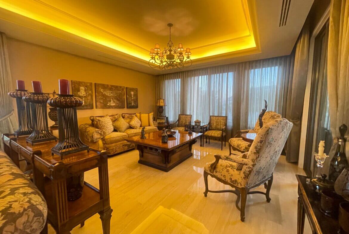 34f46589-54a0-45fb-a246-4e269010dd52 Luxurious living room with a beige upholstered sofa, floral armchairs, and a large wooden coffee table under a decorative chandelier in warm lighting.