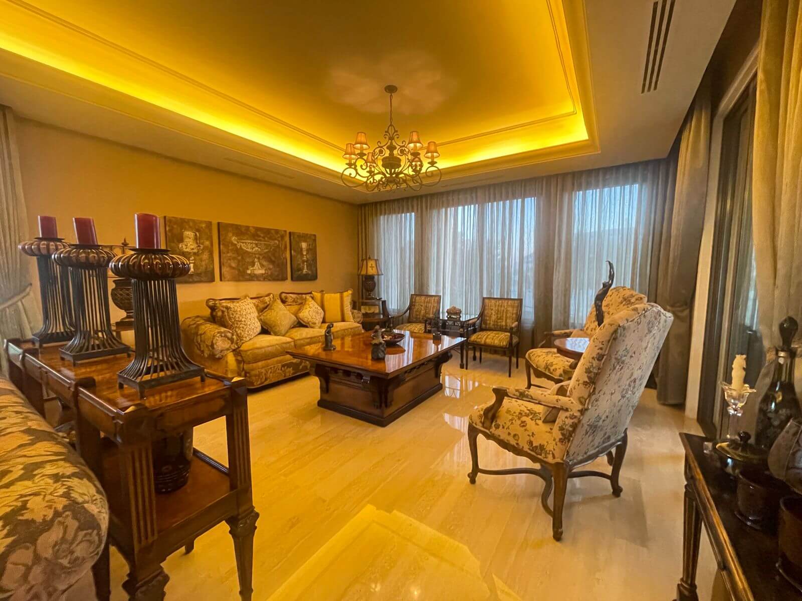 34f46589-54a0-45fb-a246-4e269010dd52 Luxurious living room with a beige upholstered sofa, floral armchairs, and a large wooden coffee table under a decorative chandelier in warm lighting.