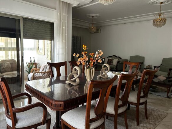 Elegant dining room with a polished wood table, upholstered chairs, decorative centerpieces, and crystal chandeliers; living area visible in the background.