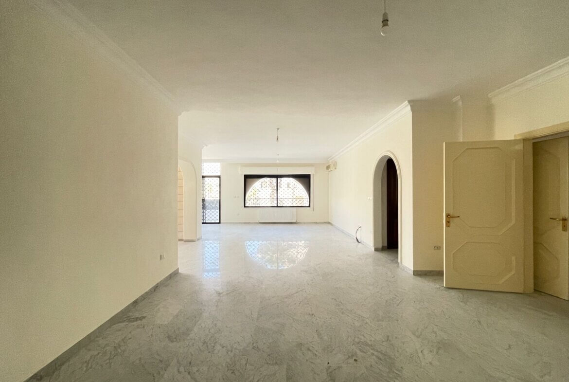 361b3204-807c-4ffe-8be2-7569af50d5ba Bright, empty living room with marble floors, arched doorway, and large patterned window letting in light. Open door on right side.