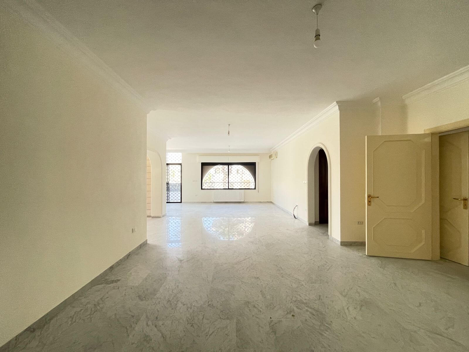 361b3204-807c-4ffe-8be2-7569af50d5ba Bright, empty living room with marble floors, arched doorway, and large patterned window letting in light. Open door on right side.