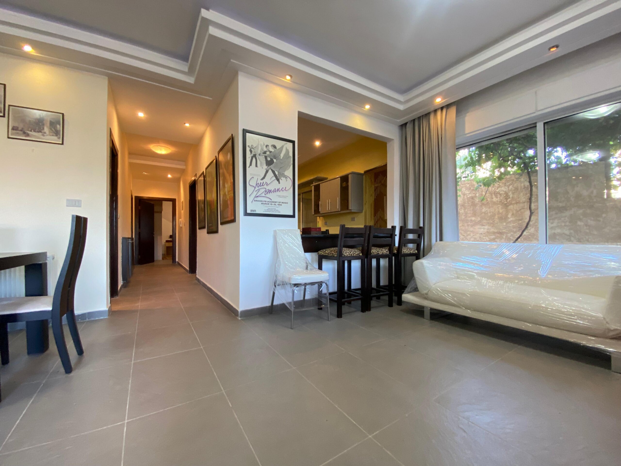 362f3d12-308c-49fd-aebe-0ec1dced64cb Open-plan living and dining area with tiled floor, wall art, and a plastic-covered sofa near large windows.