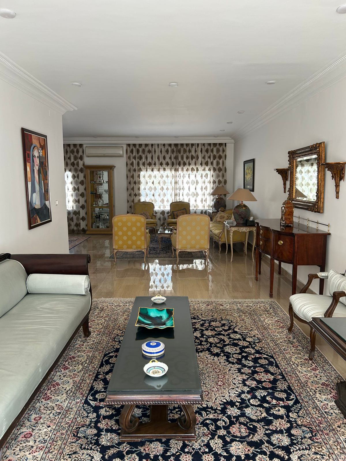 368f0740-641c-46da-ad1e-53d6305ccd71 Elegant traditional living room with a long dark coffee table on a Persian rug, yellow upholstered chairs, and antique furniture.